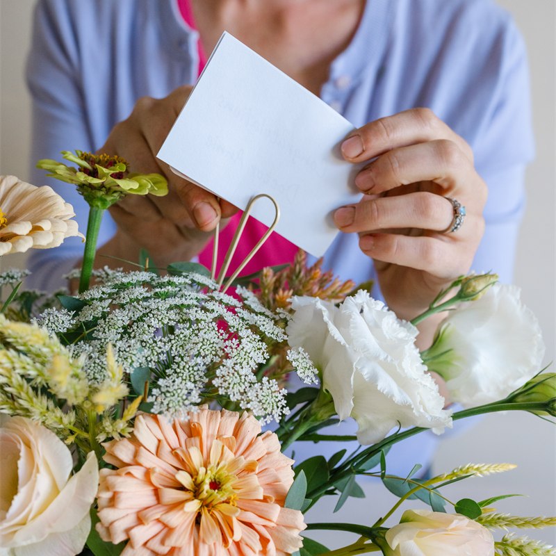 Close-up of the florist's hands placing a handwritten note into a delivery of white, yellow and pink flowers